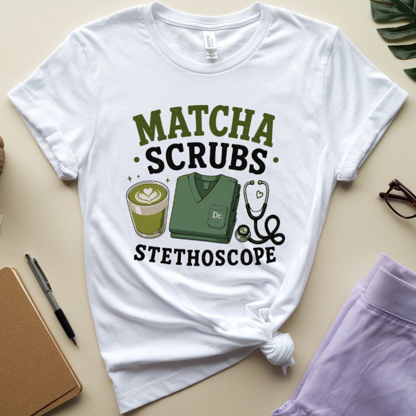 A white graphic T-shirt laid flat on a beige surface, styled with a coffee cup, notebook, pen, glasses, and a leaf. The shirt has a printed design reading “MATCHA SCRUBS STETHOSCOPE” with an illustration of a green scrub top, stethoscope, and matcha drink.