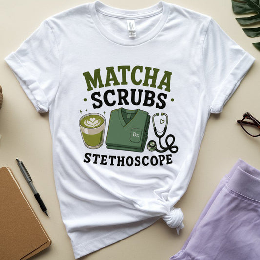 A white graphic T-shirt laid flat on a beige surface, styled with a coffee cup, notebook, pen, glasses, and a leaf. The shirt has a printed design reading “MATCHA SCRUBS STETHOSCOPE” with an illustration of a green scrub top, stethoscope, and matcha drink.