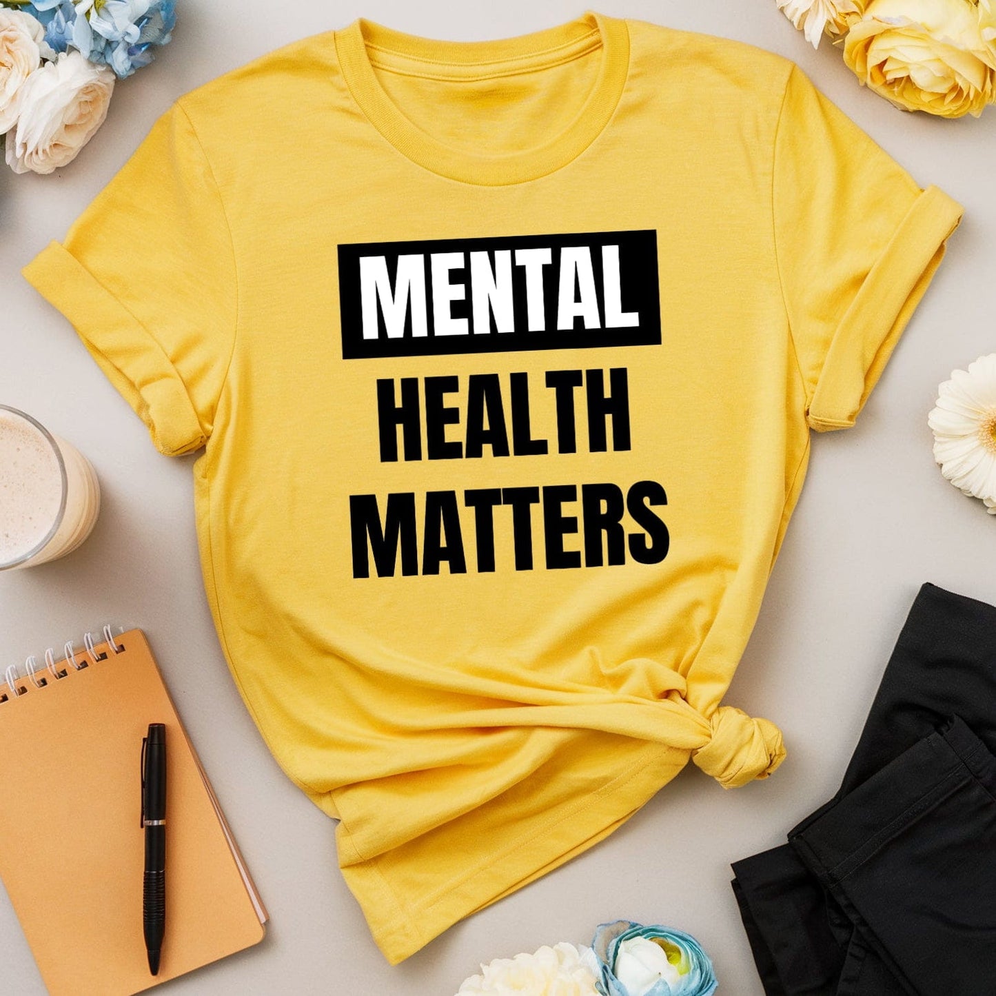 Yellow t-shirt with 'Mental Health Matters' text on a table with flowers and a smoothie.
