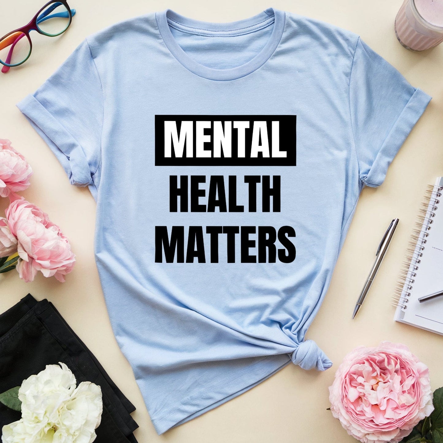 Light blue t-shirt with 'Mental Health Matters' text on a table with flowers and glasses.