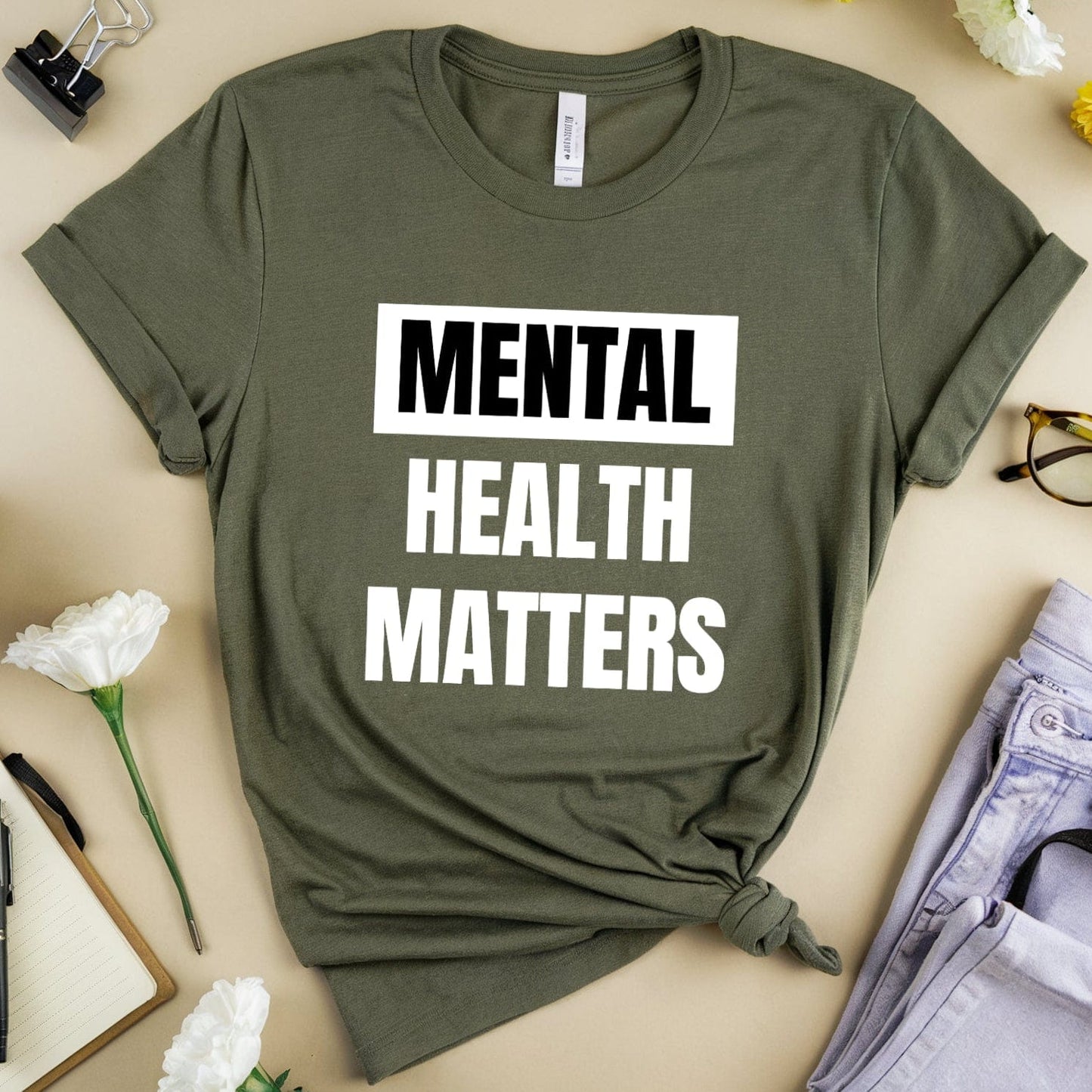 Green t-shirt with 'Mental Health Matters' text on a beige surface with flowers, coffee, and glasses.