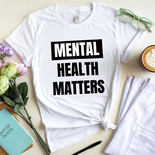White t-shirt with 'Mental Health Matters' text on a table with flowers, notebook, and coffee.