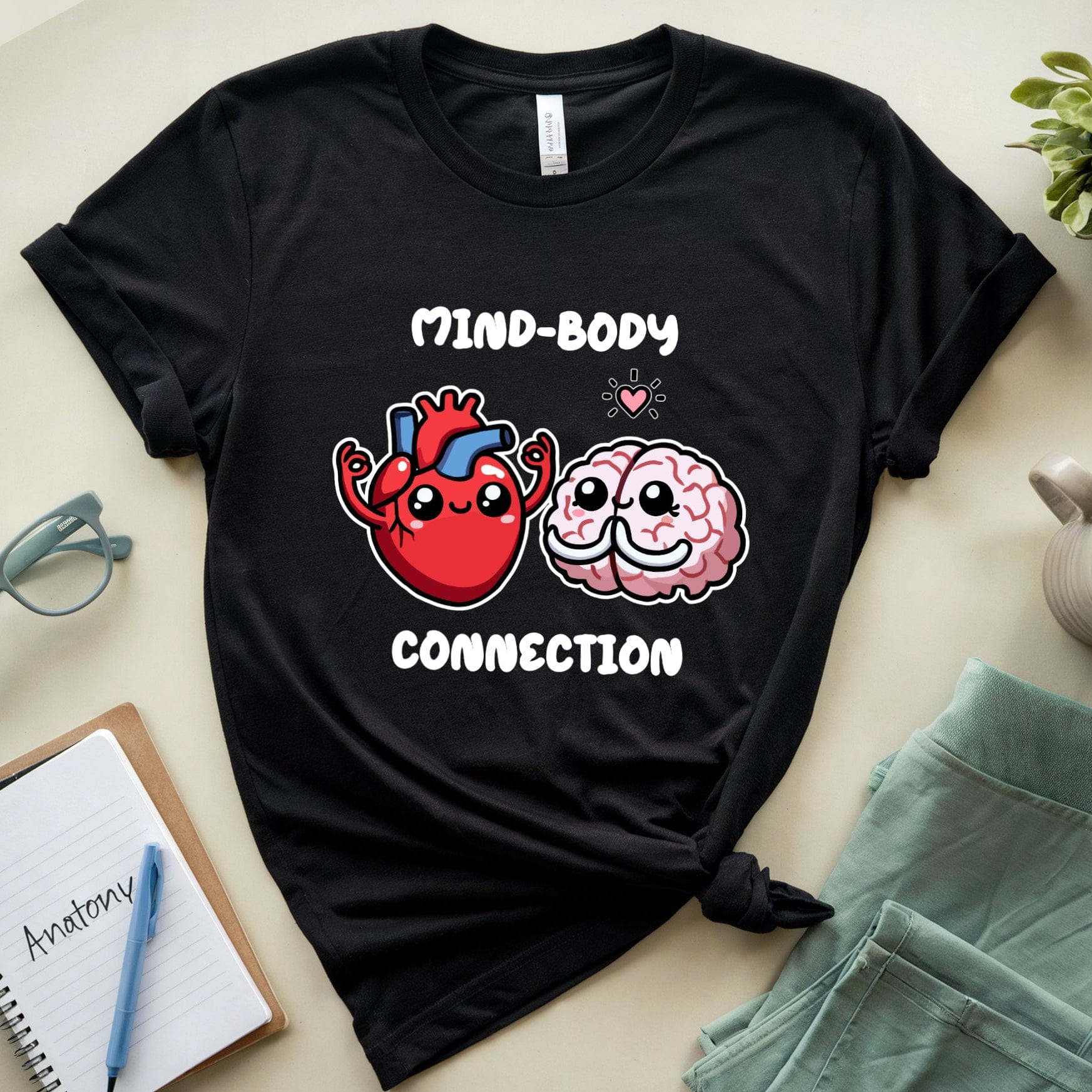 Black t-shirt with cartoon heart and brain design labeled 'Mind-Body Connection' on a light surface with accessories.