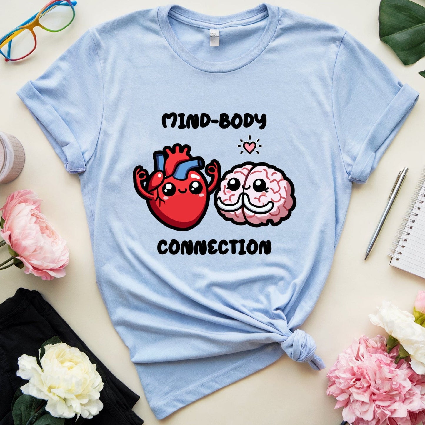 Light blue t-shirt with cartoon heart and brain design on a light surface with flowers and glasses.