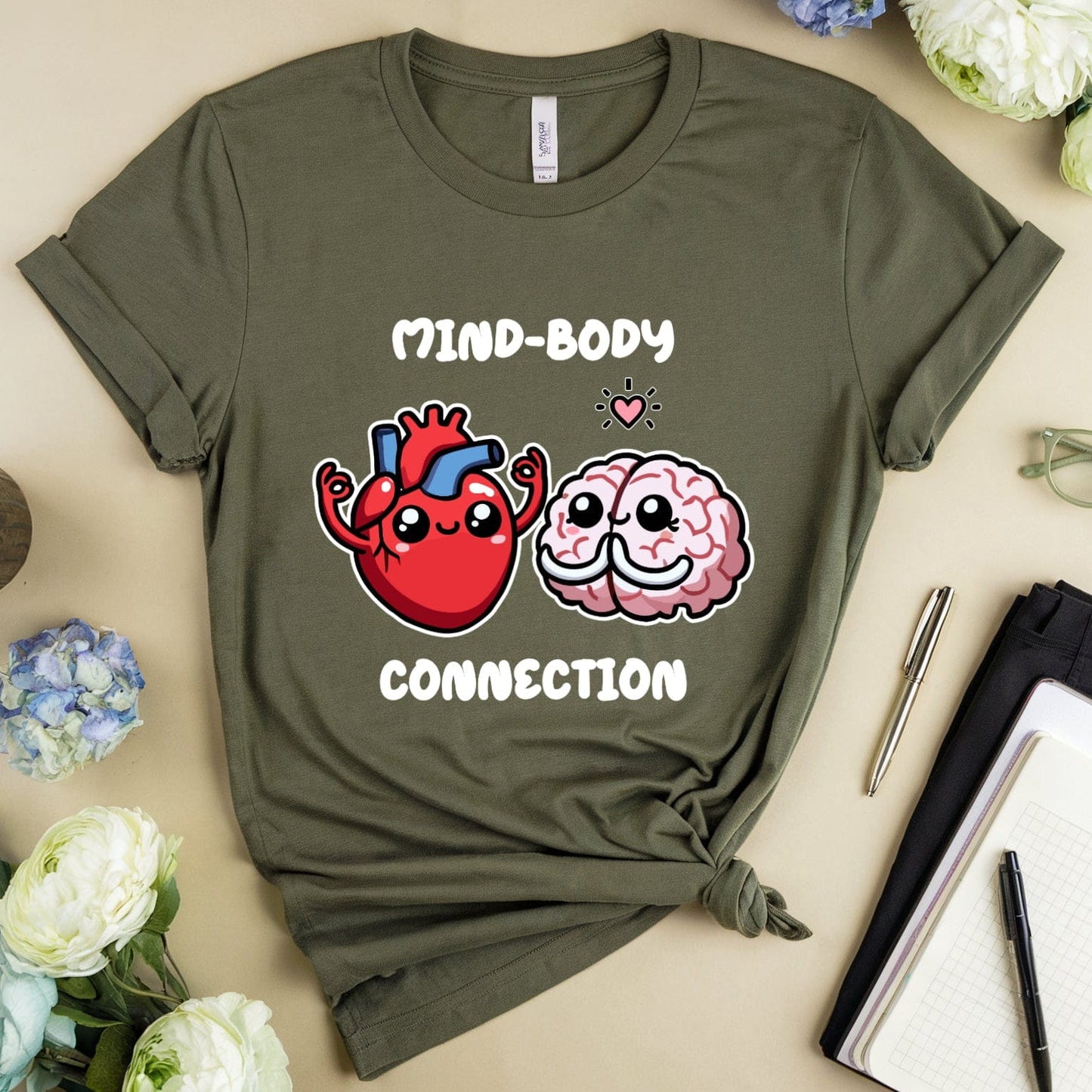 Green t-shirt with 'Mind-Body Connection' design on a beige surface with flowers and a mug.