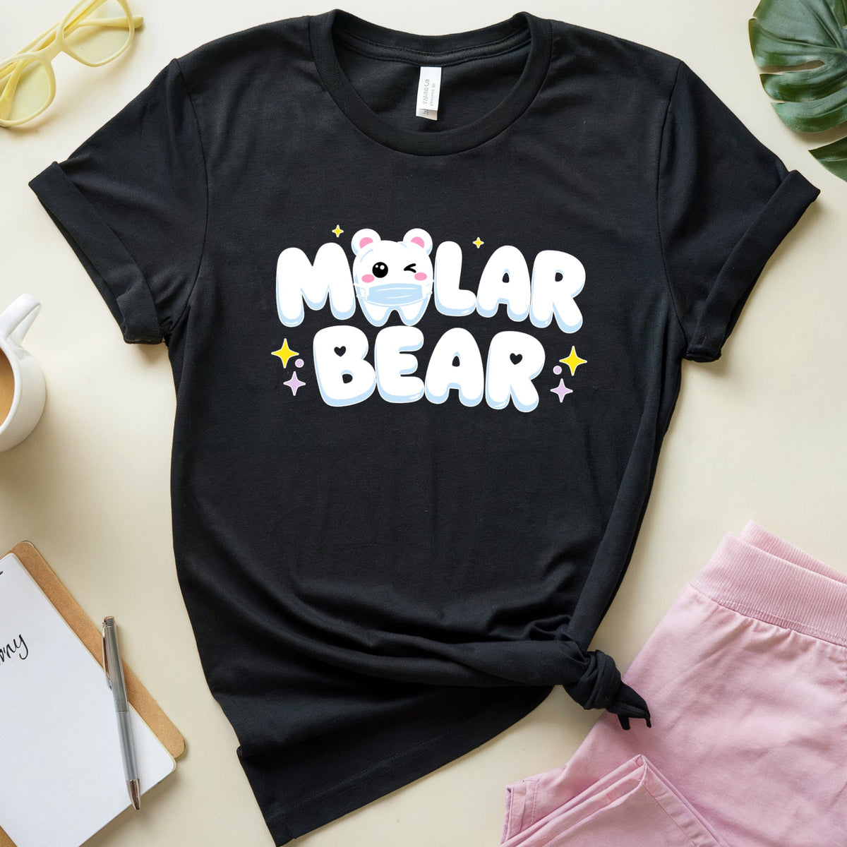 A black graphic t-shirt featuring a cute cartoon polar bear face and the text 'Molar Bear' in white with colorful star accents. The t-shirt is styled in a flat-lay arrangement on a light beige background, surrounded by pink shorts, yellow sunglasses, a coffee cup, a notebook, and a green leaf.