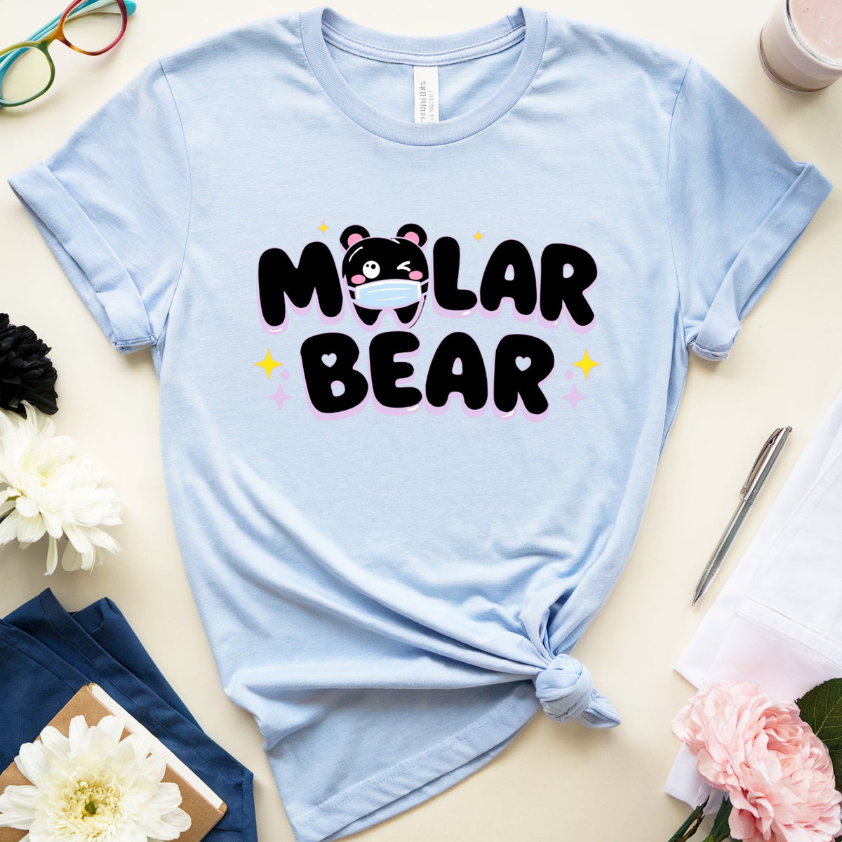 A light blue graphic t-shirt with 'MOLAR BEAR' printed in black and pink with a bear face graphic, styled in a flat lay with flowers, glasses, and a drink on a beige background.