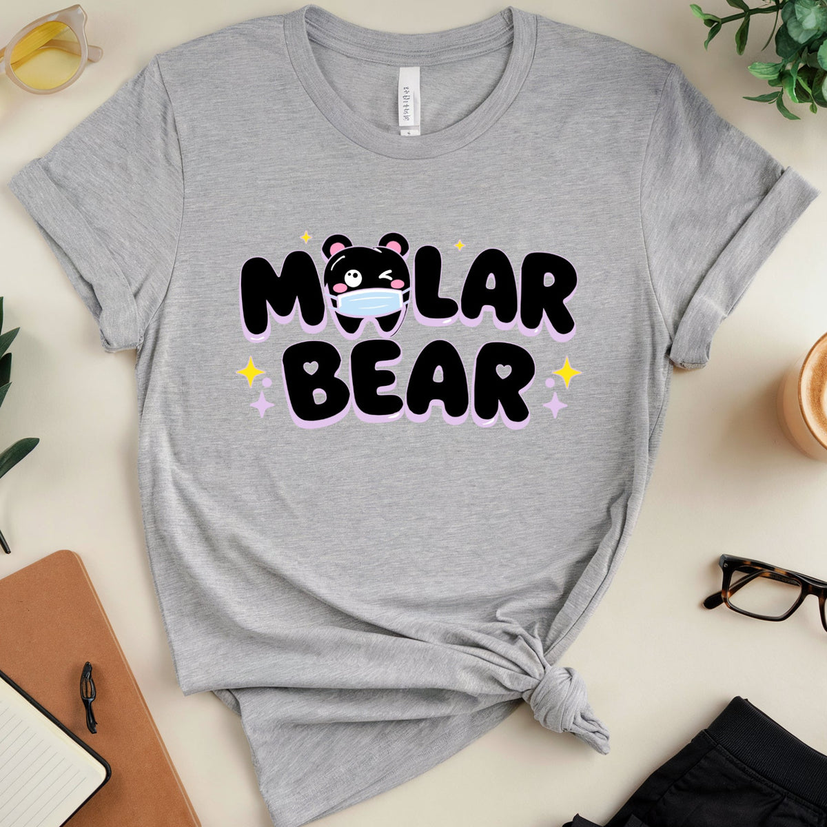 A heather gray short-sleeve t-shirt with a 'Molar Bear' graphic, styled in a flat-lay arrangement with sunglasses, a notebook, glasses, and a coffee cup on a light beige background.