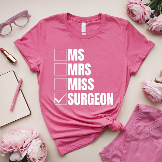Pink t-shirt with 'MS, MRS, MISS, SURGEON' text on a light surface with pink accessories.