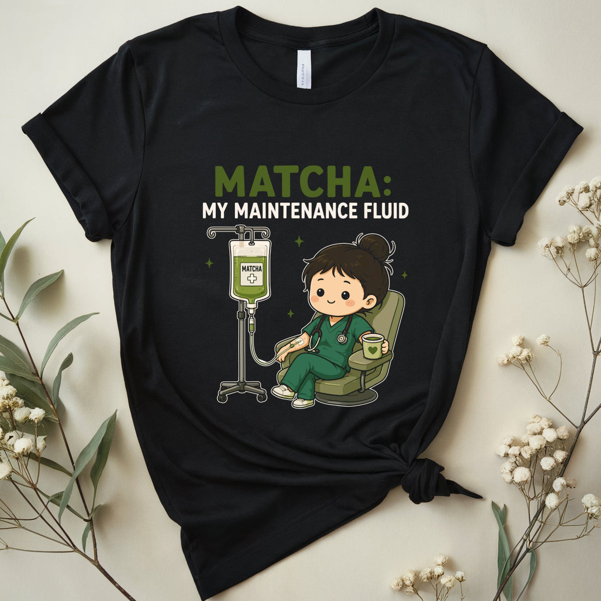 A black graphic t-shirt featuring a cartoon nurse drinking matcha and the text 'MATCHA: MY MAINTENANCE FLUID'. The t-shirt is styled in a flat-lay arrangement with dried baby's breath flowers and green leaves on a light beige background.