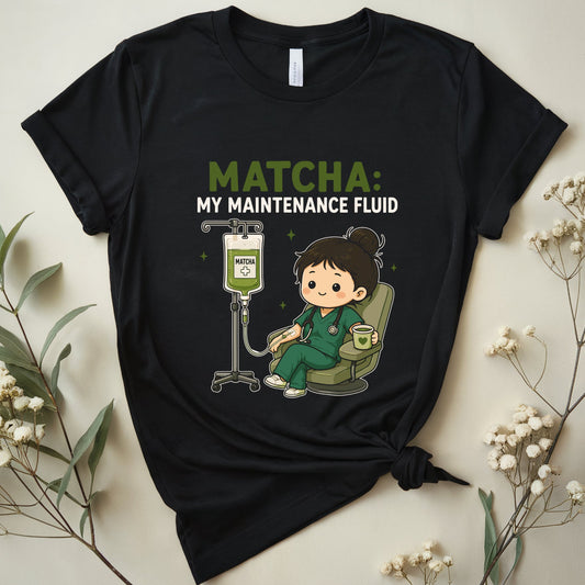 A black graphic t-shirt featuring a cartoon nurse drinking matcha and the text 'MATCHA: MY MAINTENANCE FLUID'. The t-shirt is styled in a flat-lay arrangement with dried baby's breath flowers and green leaves on a light beige background.