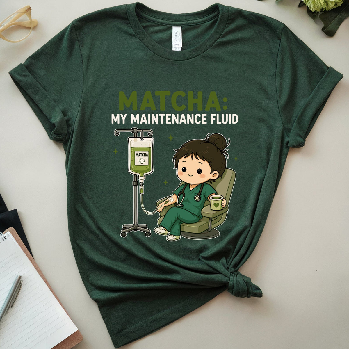 A dark green graphic t-shirt featuring a cartoon nurse and the text 'MATCHA: MY MAINTENANCE FLUID'. The t-shirt is styled in a flat-lay arrangement with a notebook, glasses, flowers, and a plant on a light beige background.