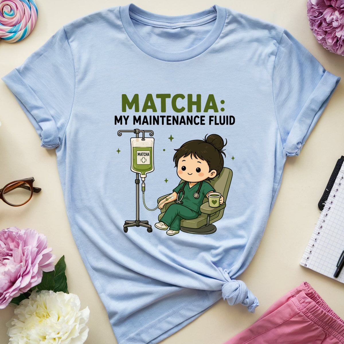 A light blue graphic t-shirt featuring a cartoon nurse sitting next to an IV bag with the text 'MATCHA: MY MAINTENANCE FLUID'. The t-shirt is styled in a flat-lay arrangement with pink and white peonies, a lollipop, eyeglasses, a notebook, and pink pants on a beige background.