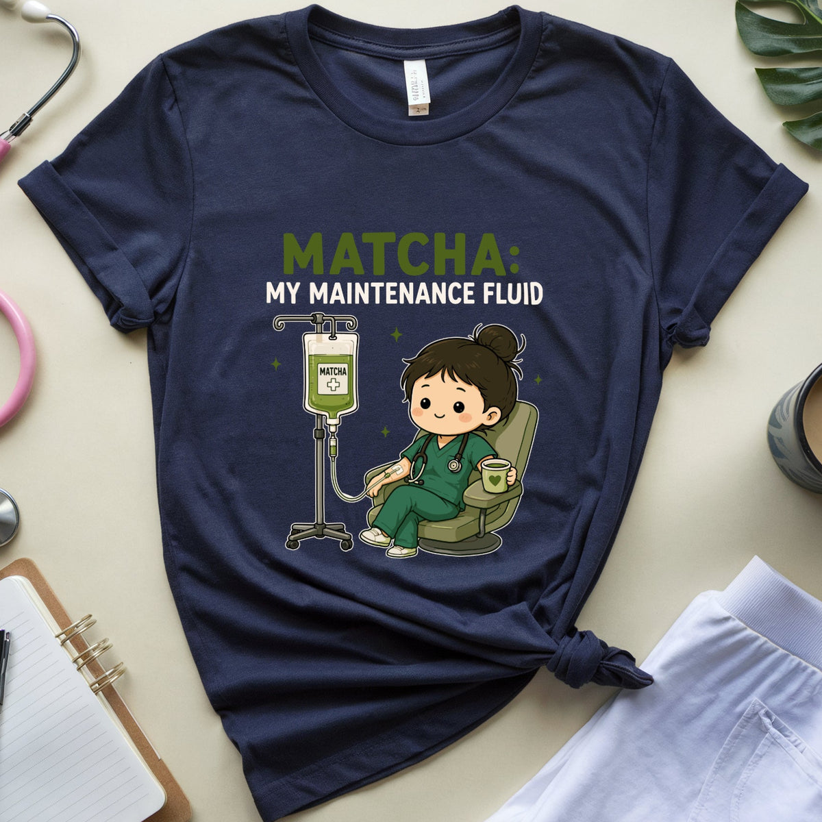 A navy blue graphic t-shirt featuring a cute cartoon nurse character with matcha hair and the text 'MATCHA MY MAINTENANCE FLUID'. The t-shirt is styled in a flat-lay arrangement with a pink stethoscope, a notebook, white pants, a coffee cup, and a green leaf on a beige background.