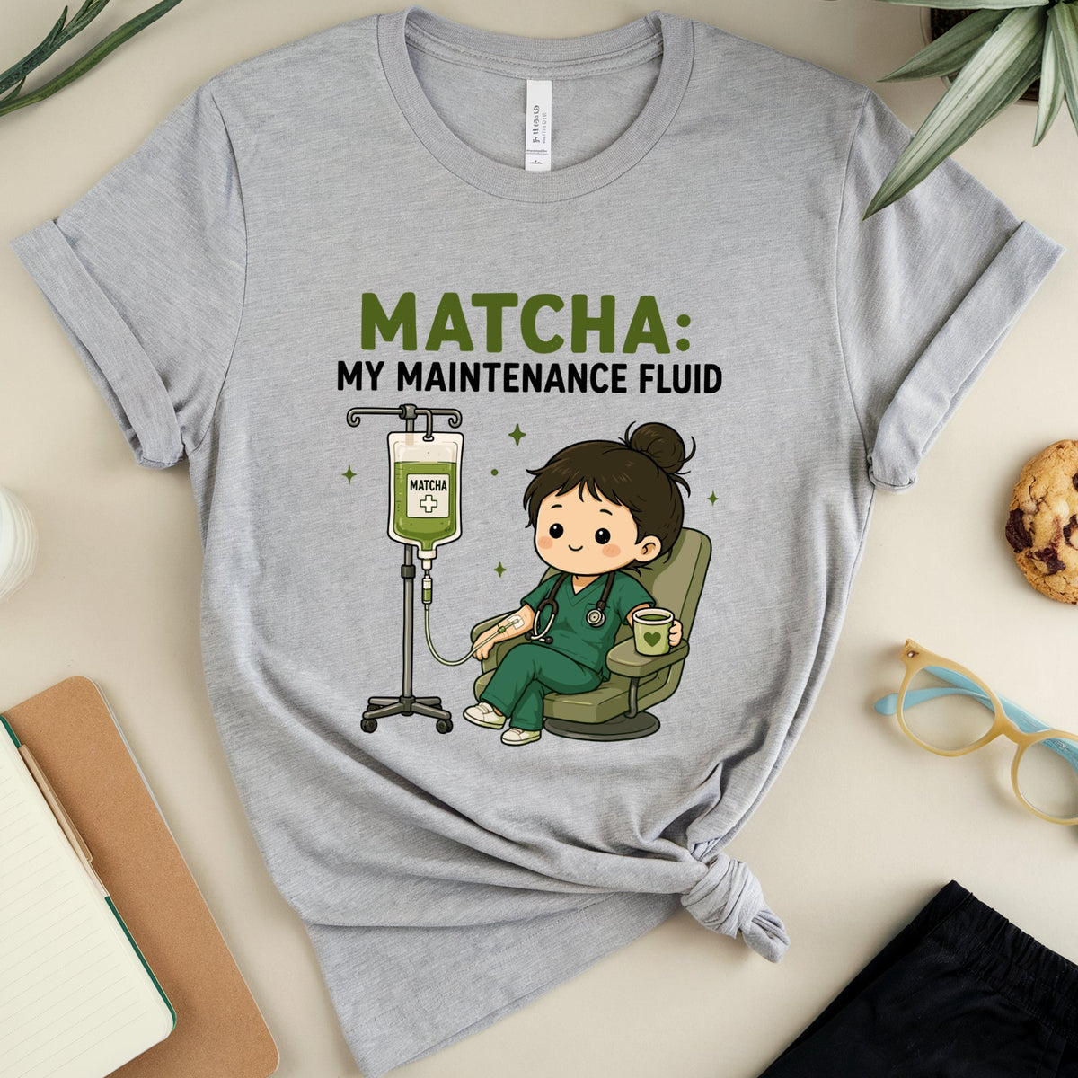 A heather gray graphic t-shirt featuring a cartoon nurse and the text 'MATCHA: MY MAINTENANCE FLUID' in green and black. The t-shirt is styled in a flat-lay arrangement on a beige surface with a plant, a notebook, a cookie, glasses, and a pair of pants.