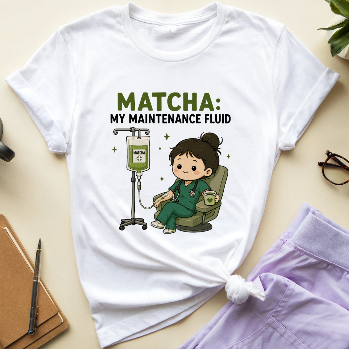 A white graphic T-shirt laid flat on a light beige surface, styled with a cup of coffee, notebook, glasses, and lavender scrubs. The shirt has a cartoon nurse sitting in an IV chair with the text 'MATCHA: MY MAINTENANCE FLUID'.