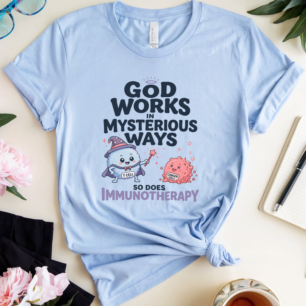 A light blue graphic t-shirt is displayed in a styled flat-lay arrangement on a cream-colored surface. The shirt features a cartoon illustration of a white cat wearing a wizard hat and staff, next to a pink monster, with the text 'GOD WORKS IN MYSTERIOUS WAYS SO DOES IMMUNOTHERAPY'.