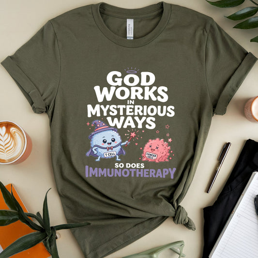 An olive green graphic t-shirt is displayed in a styled flat-lay arrangement on a beige background, surrounded by a cup of latte, notebooks, glasses, and green leaves. The t-shirt features a white and purple cartoon design with the text 'GOD WORKS IN MYSTERIOUS WAYS SO DOES IMMUNOTHERAPY'.