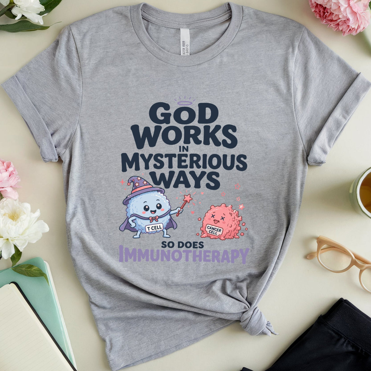 A heather gray graphic T-shirt laid flat on a light beige surface with flowers, glasses, a notebook, and a cup of tea around it. The shirt has a printed design that reads “GOD WORKS IN MYSTERIOUS WAYS SO DOES IMMUNOTHERAPY” with cartoon medical/botanical characters.