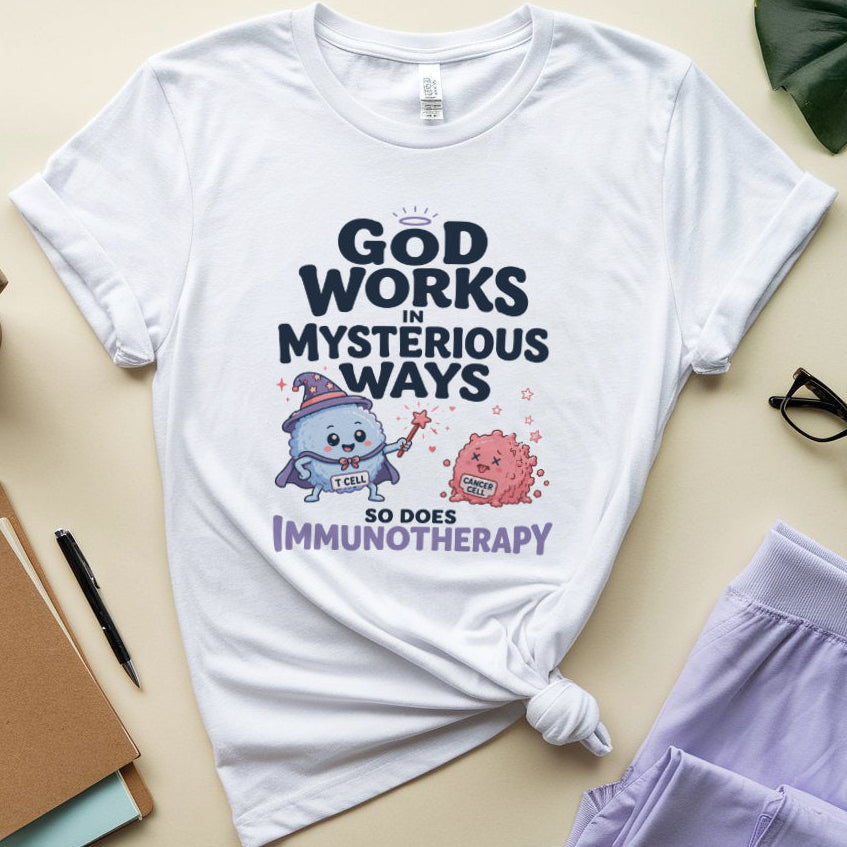 A white graphic T-shirt laid flat on a light beige surface, surrounded by lifestyle props including a notebook, pen, coffee cup, eyeglasses, and lavender pants. The shirt has a printed design with the text 'GOD WORKS IN MYSTERIOUS WAYS SO DOES IMMUNOTHERAPY' and two cartoon medical-themed characters.