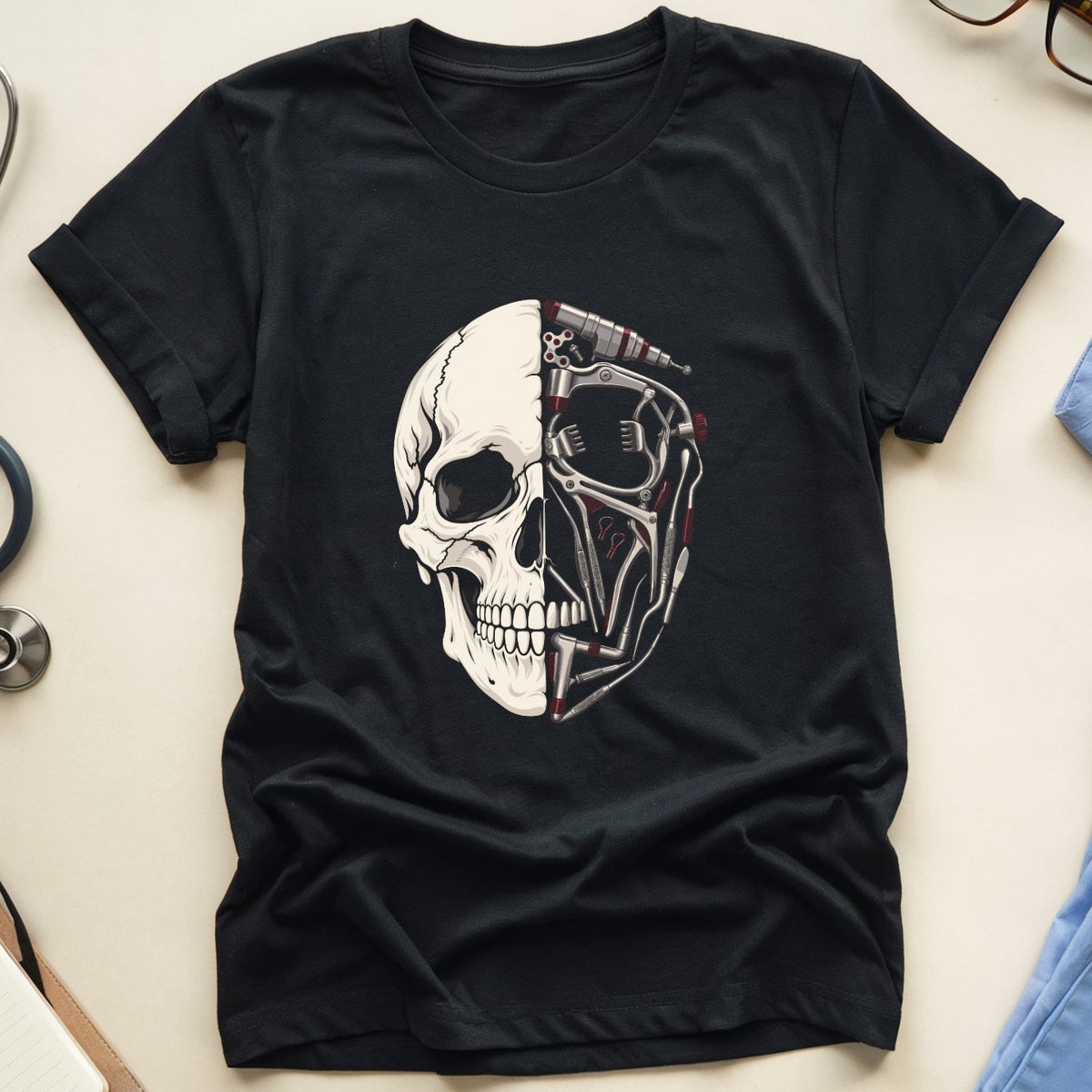 A black short-sleeve T-shirt laid flat on a light beige surface, featuring a large graphic of a split human skull in white and gray. Surrounding props include a stethoscope, eyeglasses, a notebook with a pen, and light blue medical scrubs.