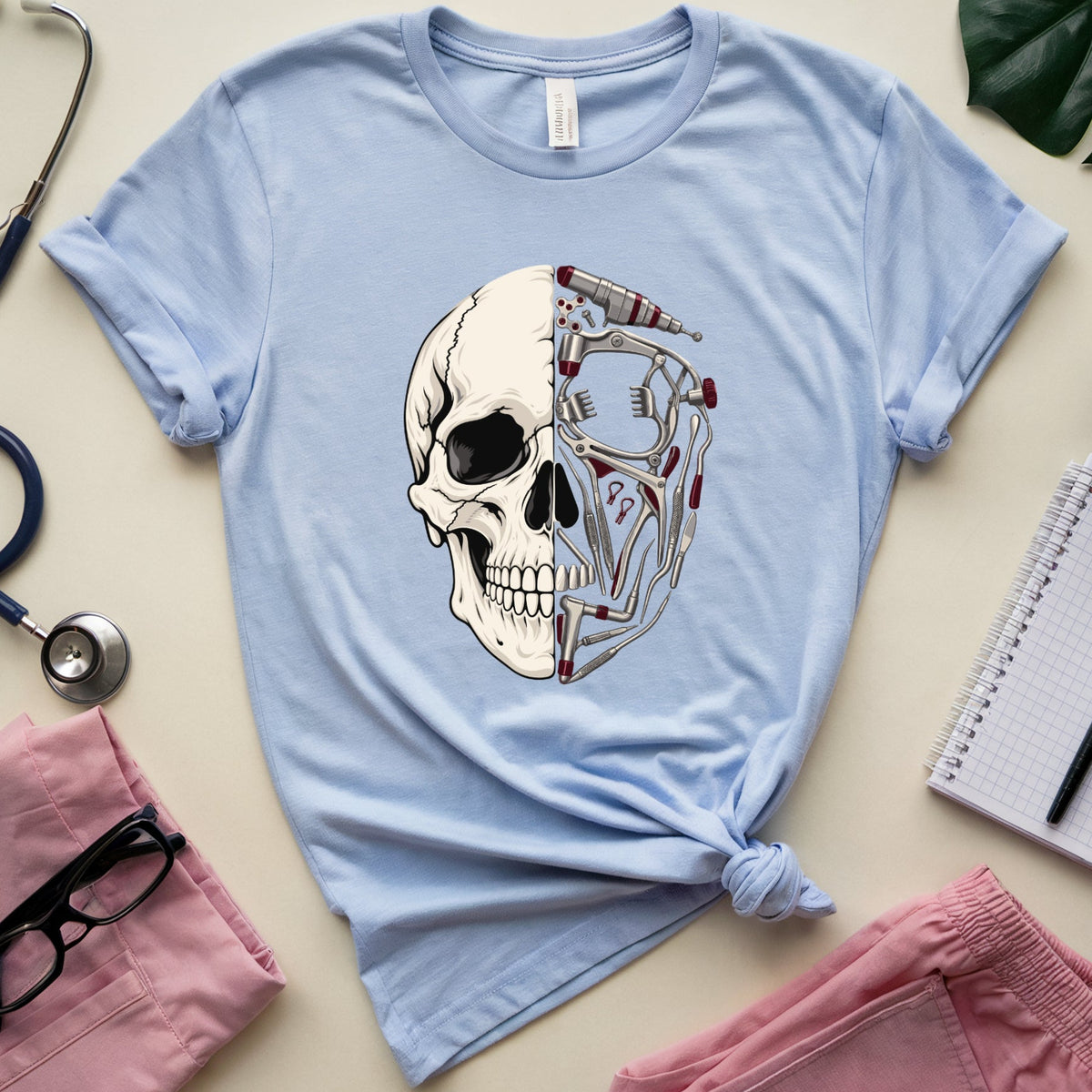 A light blue short-sleeve T-shirt laid flat on a cream surface, styled with medical-themed props including a stethoscope, pink scrubs, glasses, a notebook, and a leaf. The shirt has a large front graphic of a split human skull, one half showing a normal skull and the other half showing exposed bones and internal structures.
