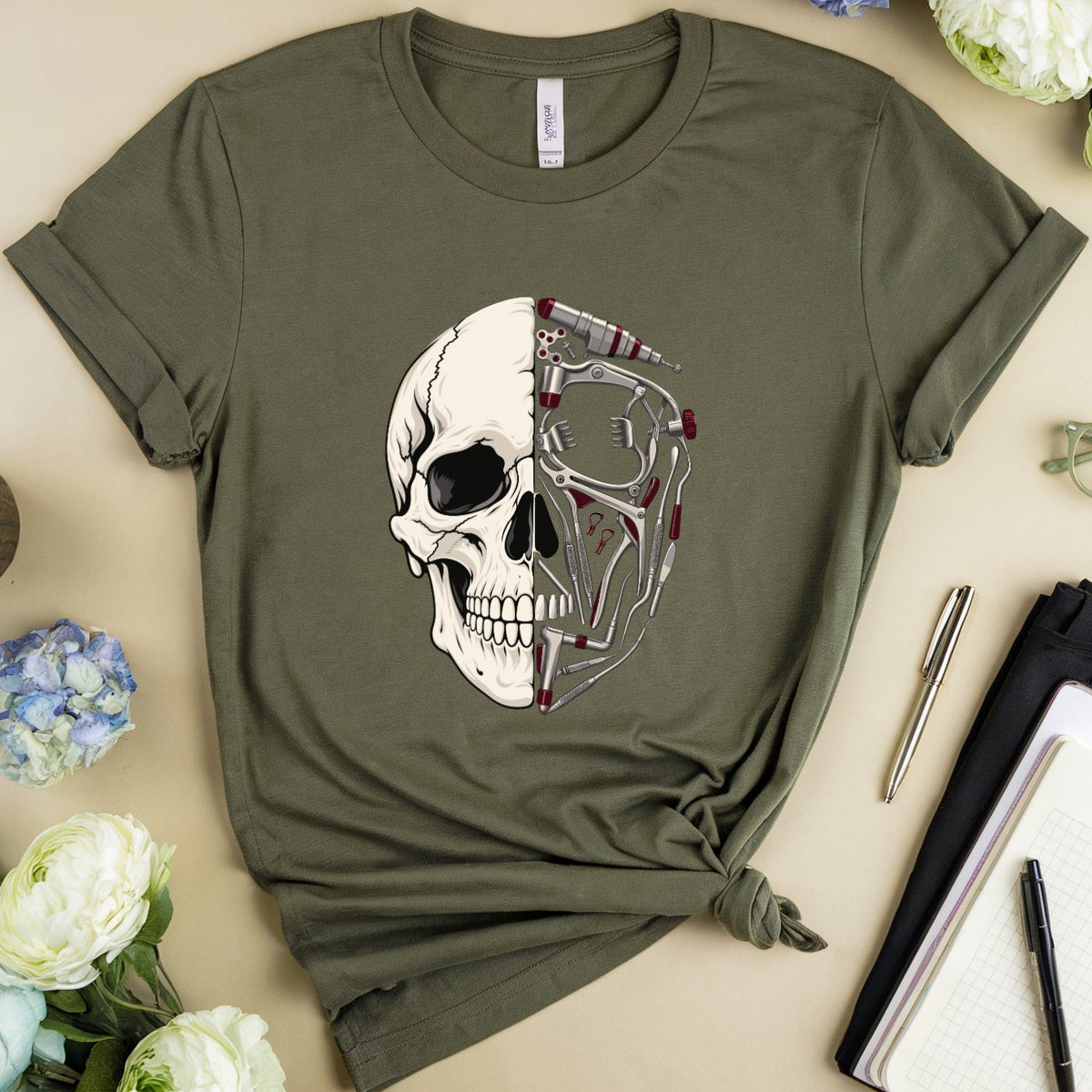 An olive green short-sleeve t-shirt with a graphic of a skull and glasses, styled in a flat lay with flowers, a notebook, glasses, and a mug on a beige background.