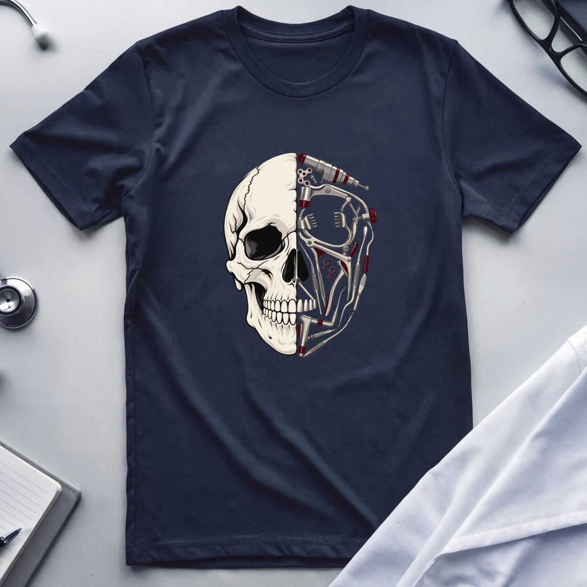 A navy blue short-sleeve t-shirt with a graphic of a split human skull, one half showing a normal skull and the other half showing an anatomical model, laid flat on a light gray surface with medical props like a stethoscope, glasses, and a lab coat.