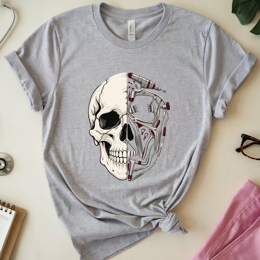A heather gray short-sleeve t-shirt with a graphic of a human skull and anatomical skeleton detailing, styled in a flat lay with medical props including a stethoscope, notebook, glasses, and pink scrubs on a light beige background.