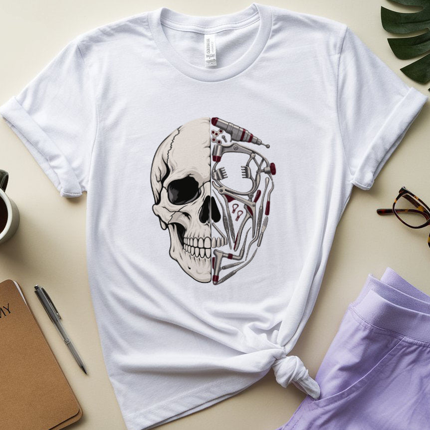A white short-sleeve graphic T-shirt laid flat on a light beige surface, styled with a coffee cup, notebook, pen, glasses, a purple garment, and a green leaf. The shirt has a large front graphic of a skull split into anatomical bone and mechanical parts with red accents.