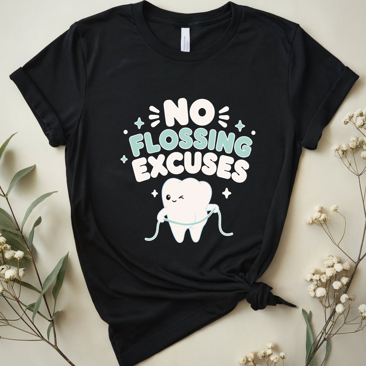 A black short-sleeve graphic T-shirt laid flat on a light beige background with dried flowers around it. The shirt has a front graphic that reads “NO FLOSSING EXCUSES” in white and mint green lettering, with a cartoon tooth holding a flossing cord and small sparkles.