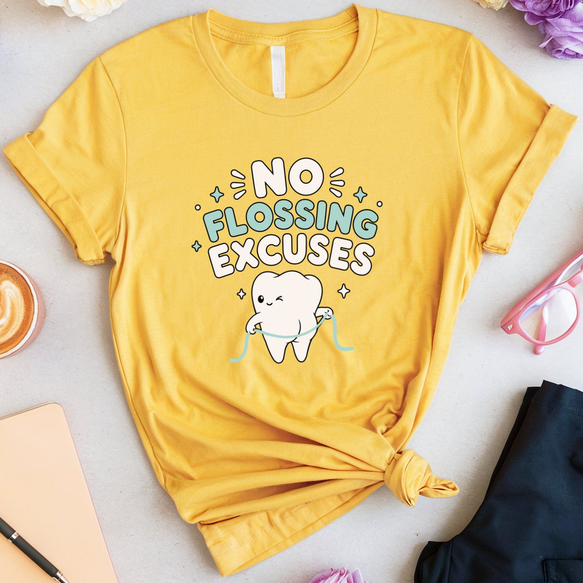 A yellow graphic T-shirt laid flat on a light surface with flowers, a coffee cup, notebook, pen, pink glasses, and dark pants around it. The shirt has a printed message reading 'NO FLOSSING EXCUSES' with a cartoon tooth and colorful decorative accents.