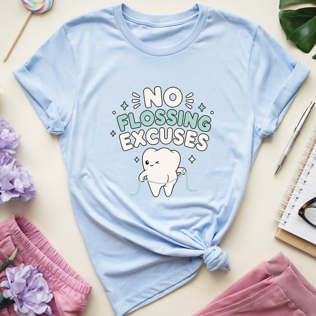 A light blue graphic t-shirt with rolled sleeves, featuring a cartoon tooth and the text 'NO FLOSSING EXCUSES'. The t-shirt is styled in a flat lay on a beige background with pink shorts, purple flowers, a lollipop, a pen, a notebook, and eyeglasses.
