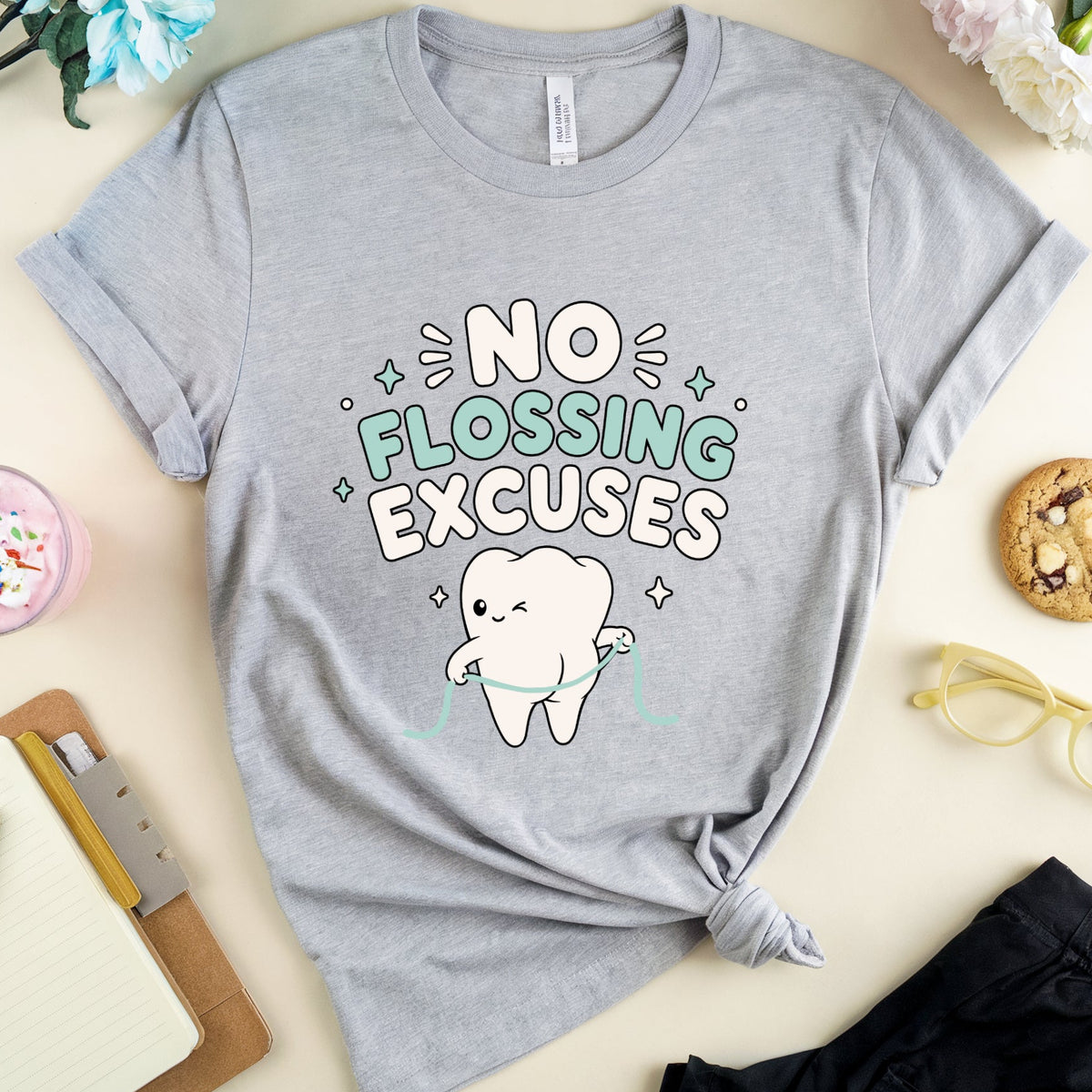 A heather gray graphic t-shirt featuring a cartoon tooth and the text 'NO FLOSSING EXCUSES' in white, black, and mint green. The shirt is styled in a flat lay on a light beige background with a pink drink, notebook, cookie, glasses, and flowers.