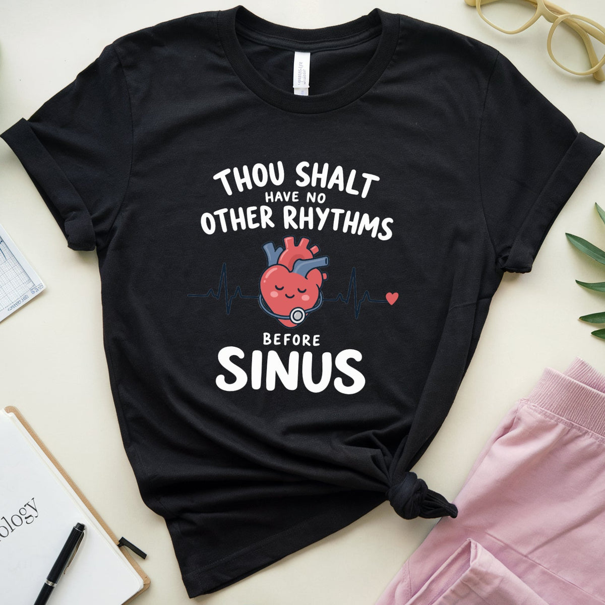 A black graphic t-shirt with a heart-shaped EKG and the text 'THOU SHALT HAVE NO OTHER RHYTHMS BEFORE SINUS' is styled in a flat lay with pink pants, green leaves, glasses, and notebooks on a light beige background.