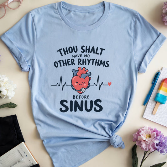 A light blue graphic t-shirt with a knotted hem, featuring a cartoon heart and EKG line with the text 'THOU SHALT HAVE NO OTHER RHYTHMS BEFORE SINUS'. The t-shirt is styled in a flat lay with glasses, flowers, a notebook, a popsicle, and pants on a beige background.