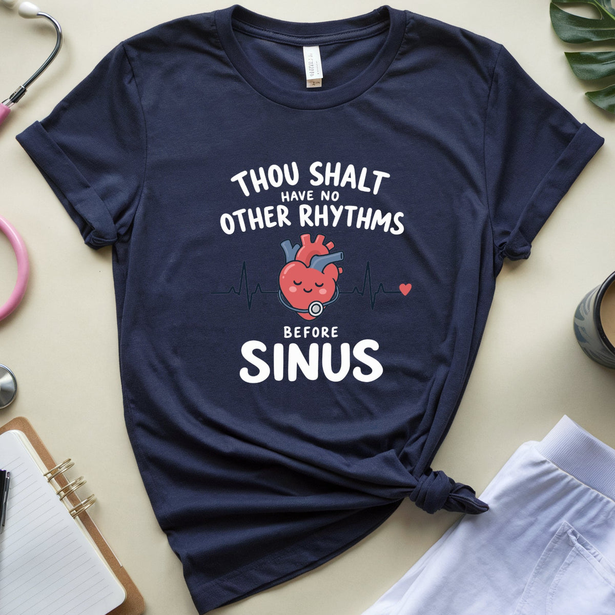 A navy blue graphic t-shirt with a medical-themed print reading 'THOU SHALT HAVE NO OTHER RHYTHMS BEFORE SINUS' and an illustration of a smiling red heart, styled in a flat lay with a pink stethoscope, notebook, coffee cup, and white pants on a beige background.
