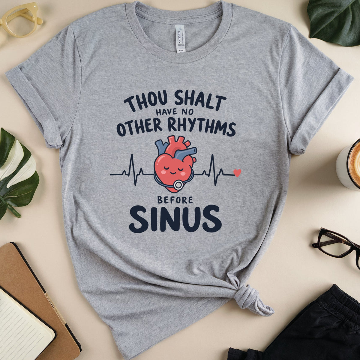 A heather gray graphic t-shirt with a 'Thou Shalt Have No Other Rhythms Before Sinus' design featuring a cartoon heart and EKG line, styled in a flat lay with glasses, a coffee cup, and notebook.