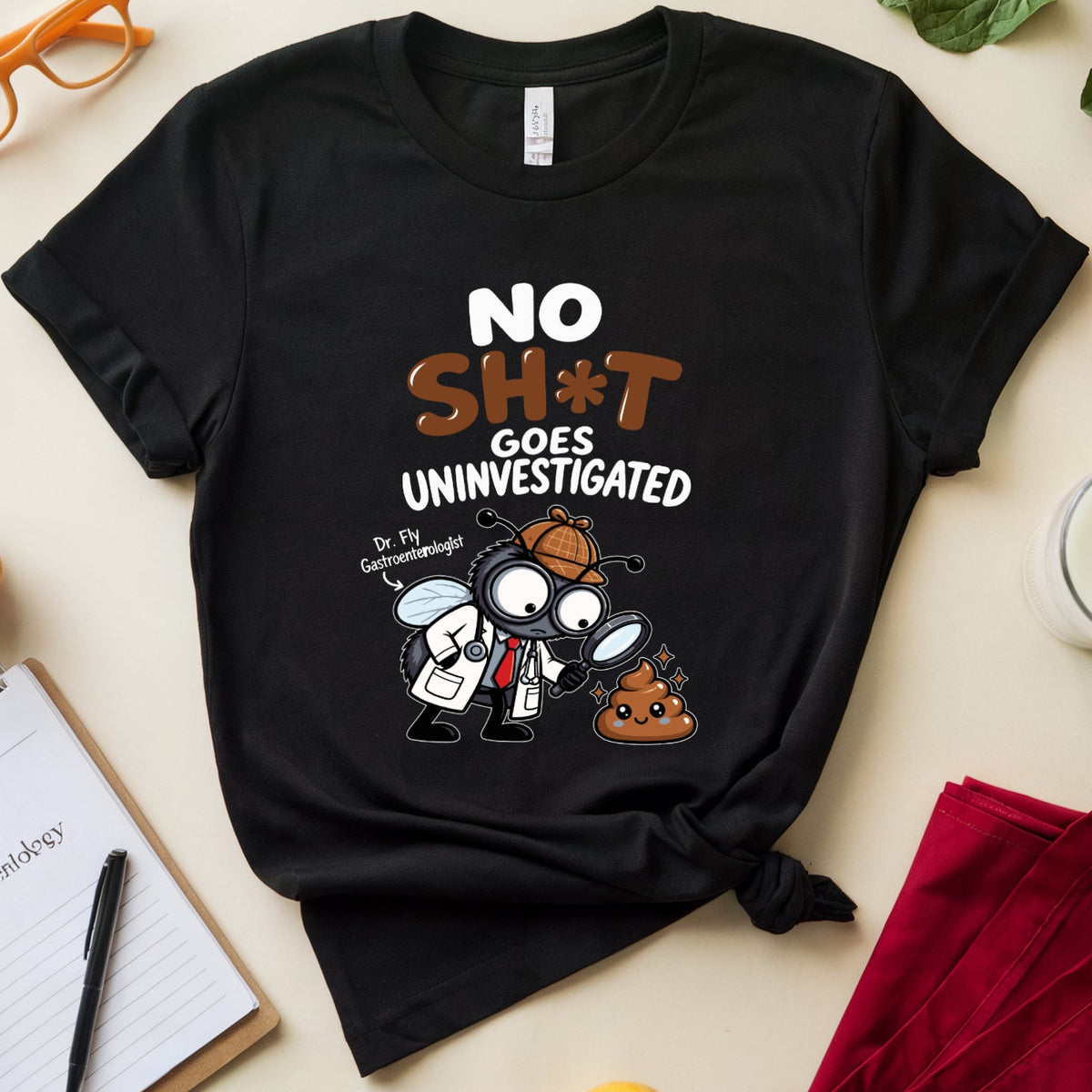 A black graphic T-shirt laid flat on a light beige surface, surrounded by styled props including orange glasses, a notebook with pen, a glass of milk, red pants, and fruits and greens. The shirt has a cartoon graphic of a bug character with magnifying glasses and the text 'NO SH*T GOES UNINVESTIGATED.