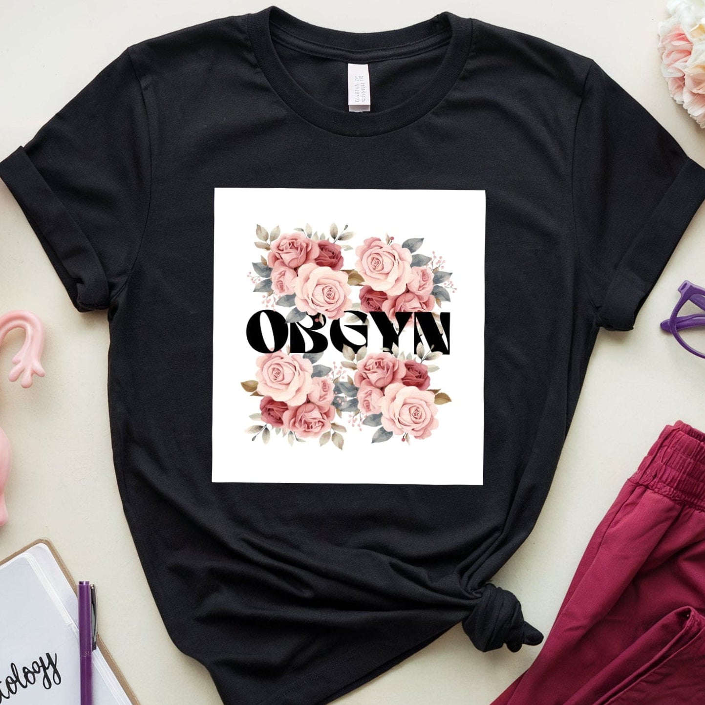 Black t-shirt with floral design and text, placed on a light surface with pink pants, notebook, and glasses.