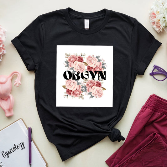 Black t-shirt with floral design and text, placed on a light surface with pink pants, notebook, and glasses.