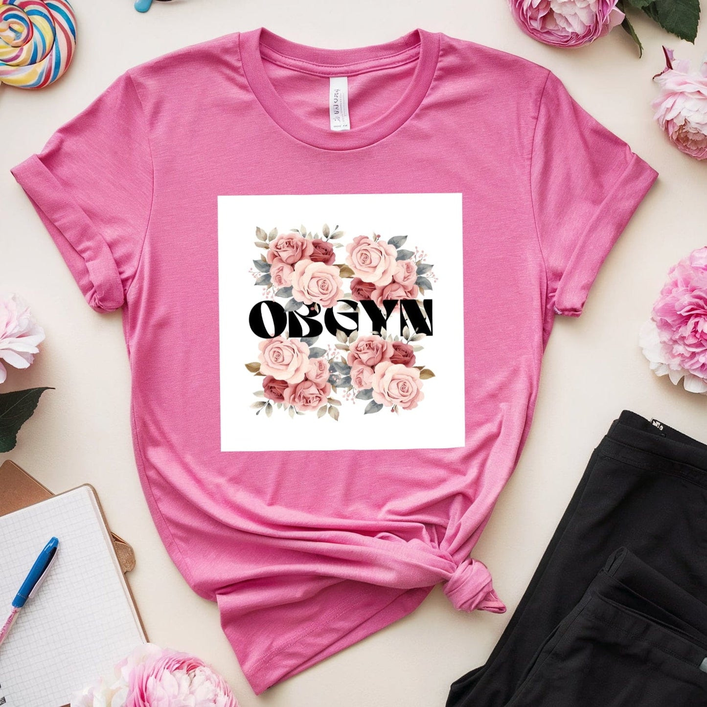 Pink t-shirt with floral design and 'OBGYN' text, surrounded by flowers and stationery on a light surface.