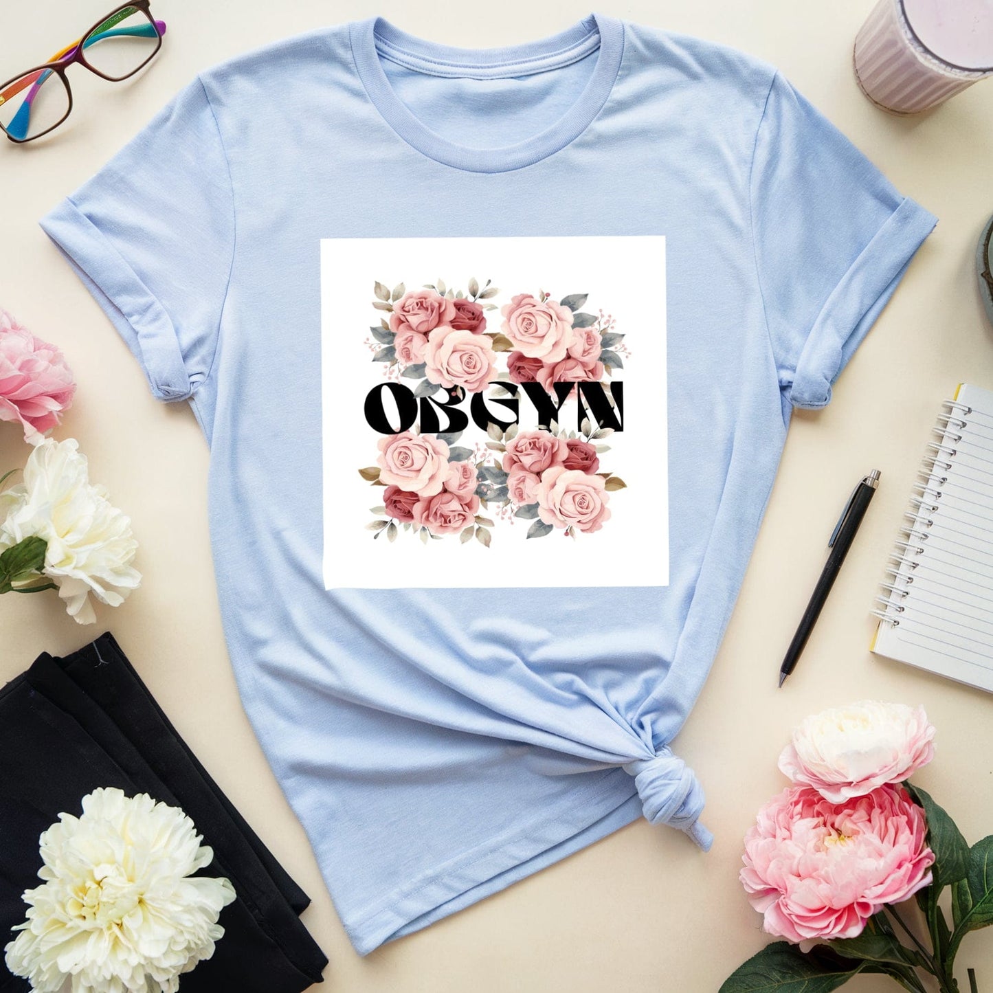 Light blue t-shirt with floral design and text on a table with flowers and glasses.