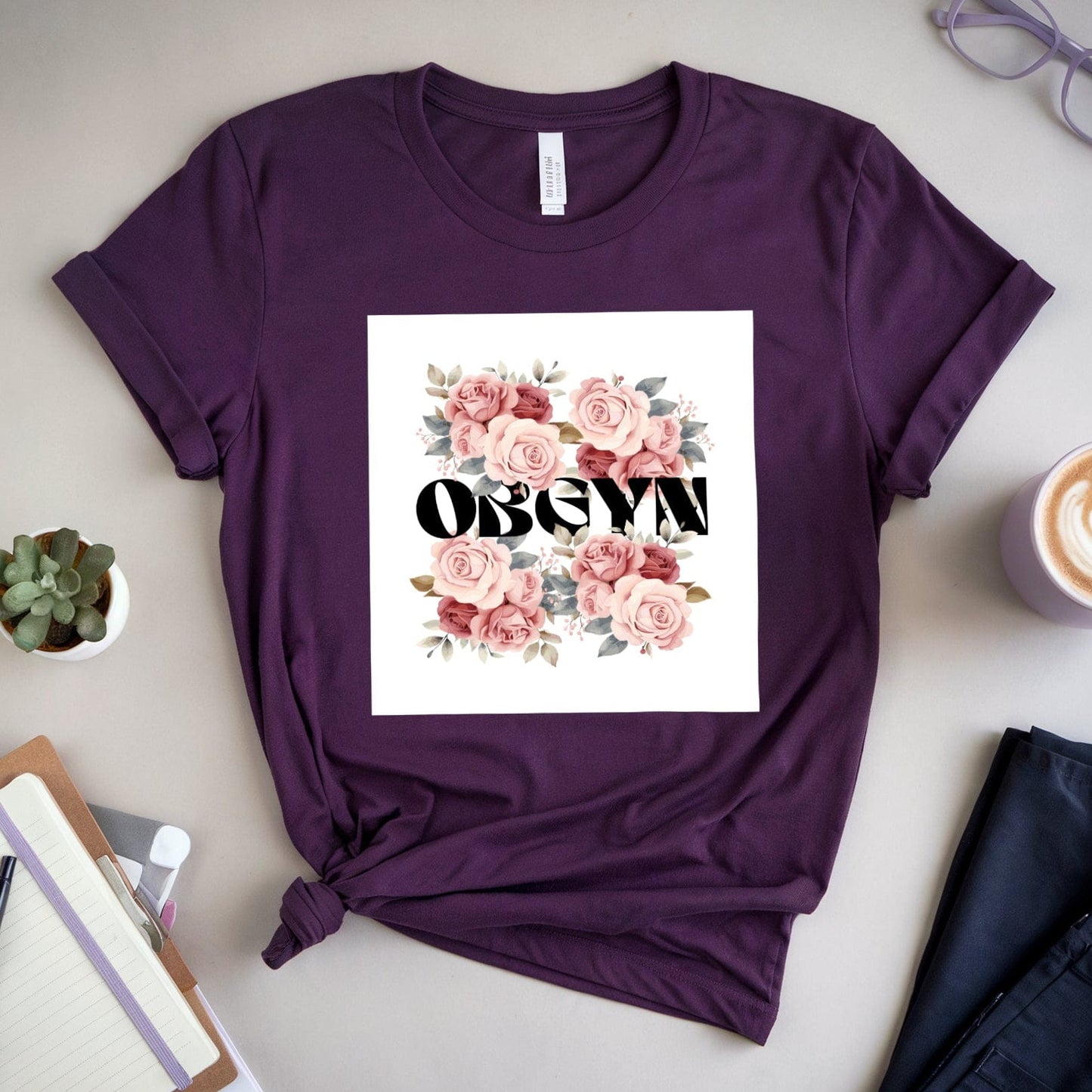 Purple t-shirt with floral design and text on a table with a cup of coffee, glasses, and notebook.