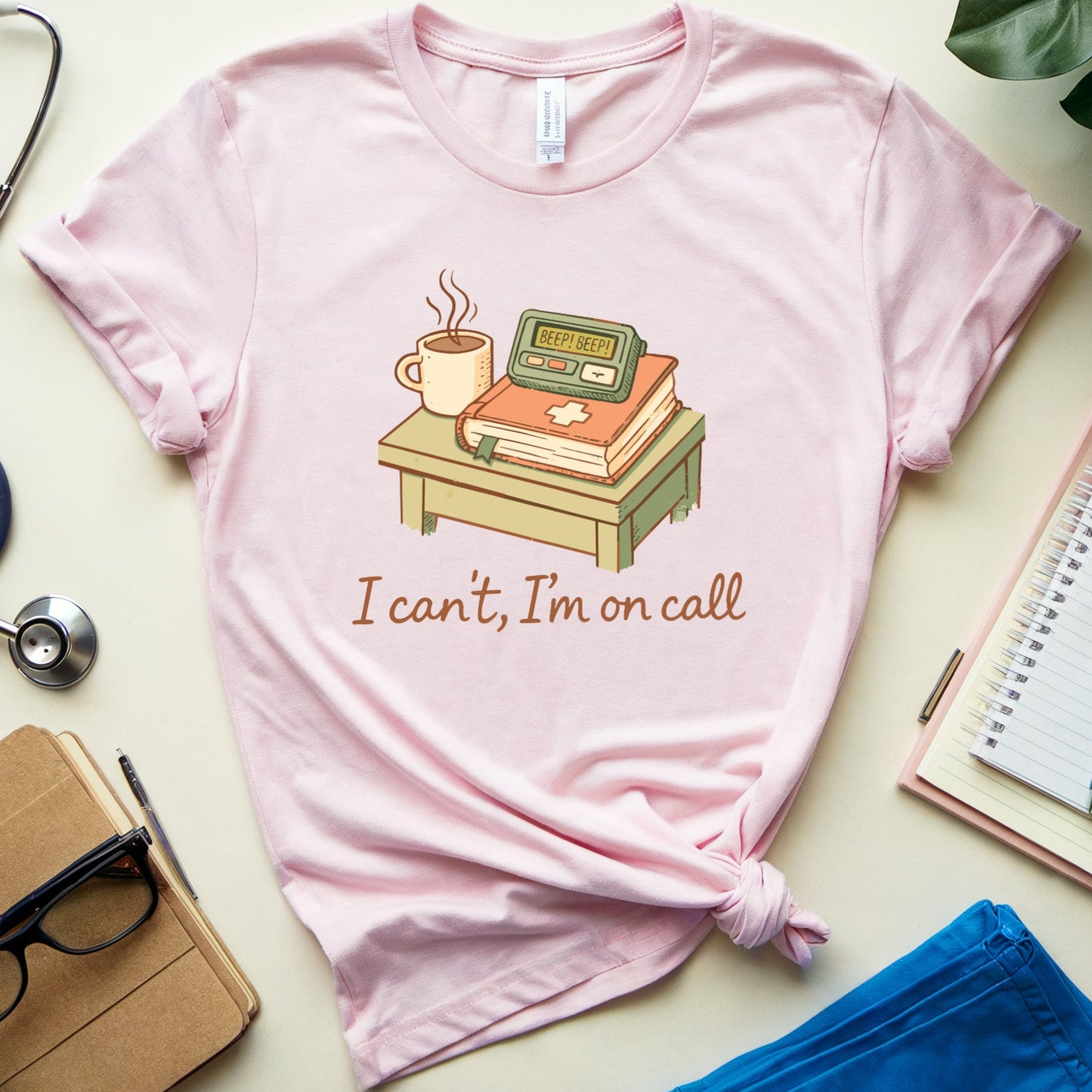 Pink t-shirt with a graphic and text 'I can't, I'm on call' on a light surface with medical-themed items.