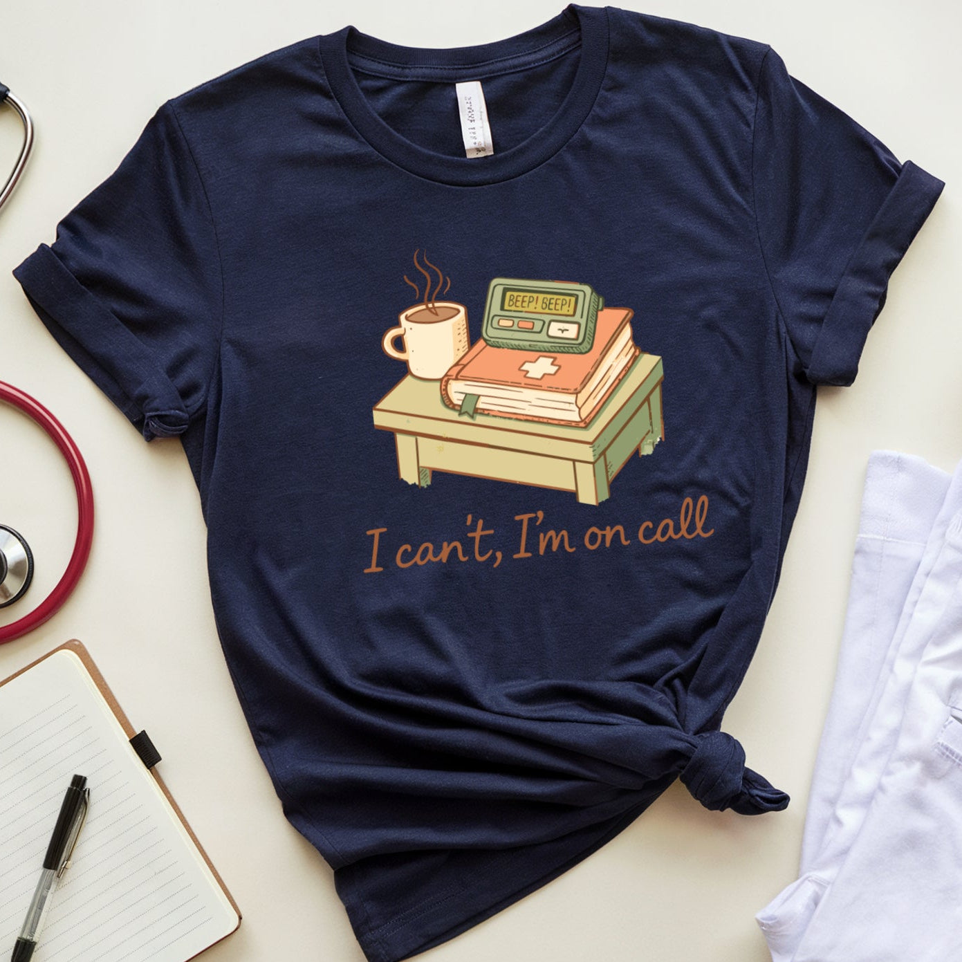 Navy blue t-shirt with a graphic and text 'I can't, I'm on call' on a light surface with medical-themed items.