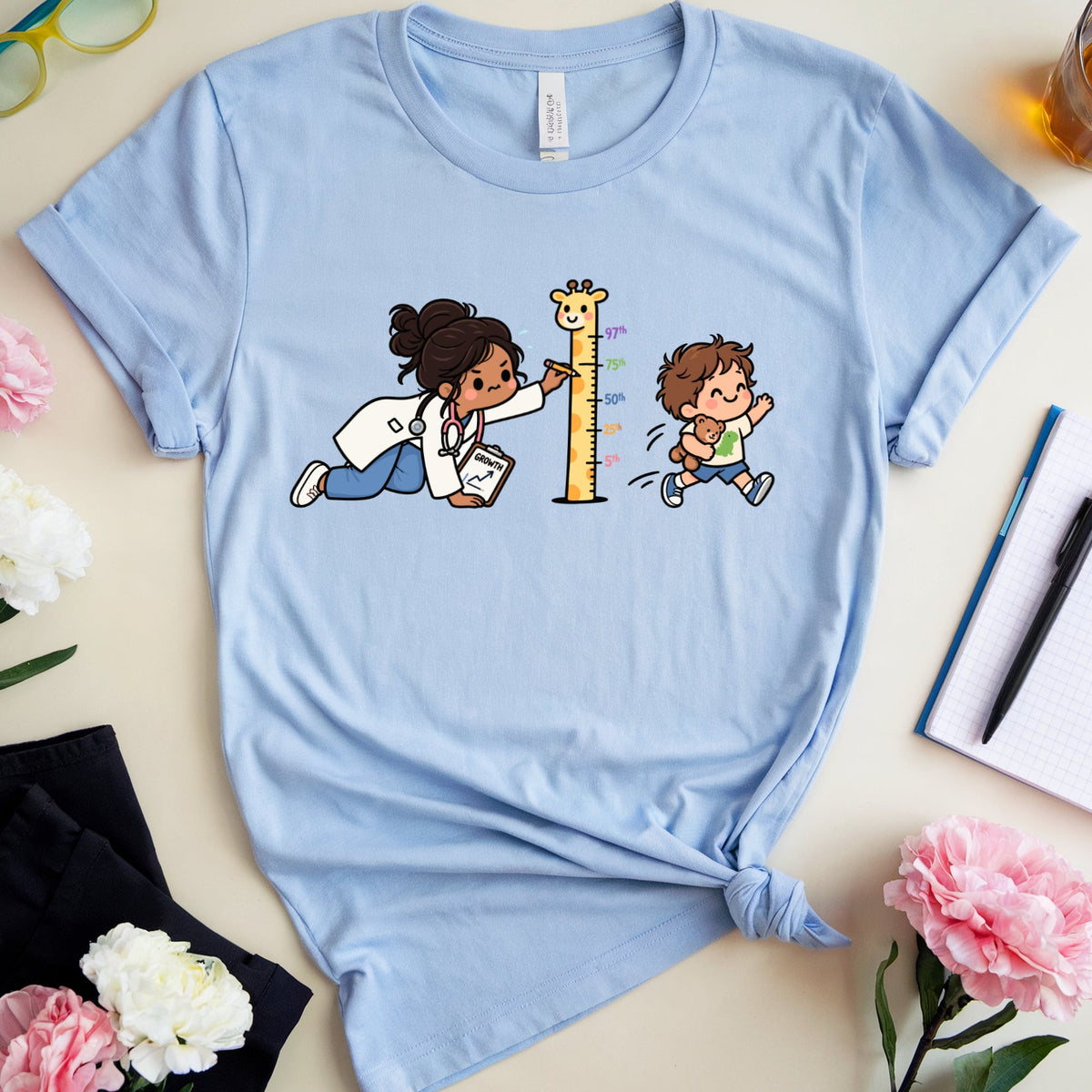A light blue graphic t-shirt laid flat on a beige surface, surrounded by decorative props including pink and white flowers, a pair of green-framed glasses, a notebook with a pen, and a glass of iced tea. The t-shirt features a cartoon graphic of a woman measuring a child's height with a tall giraffe.