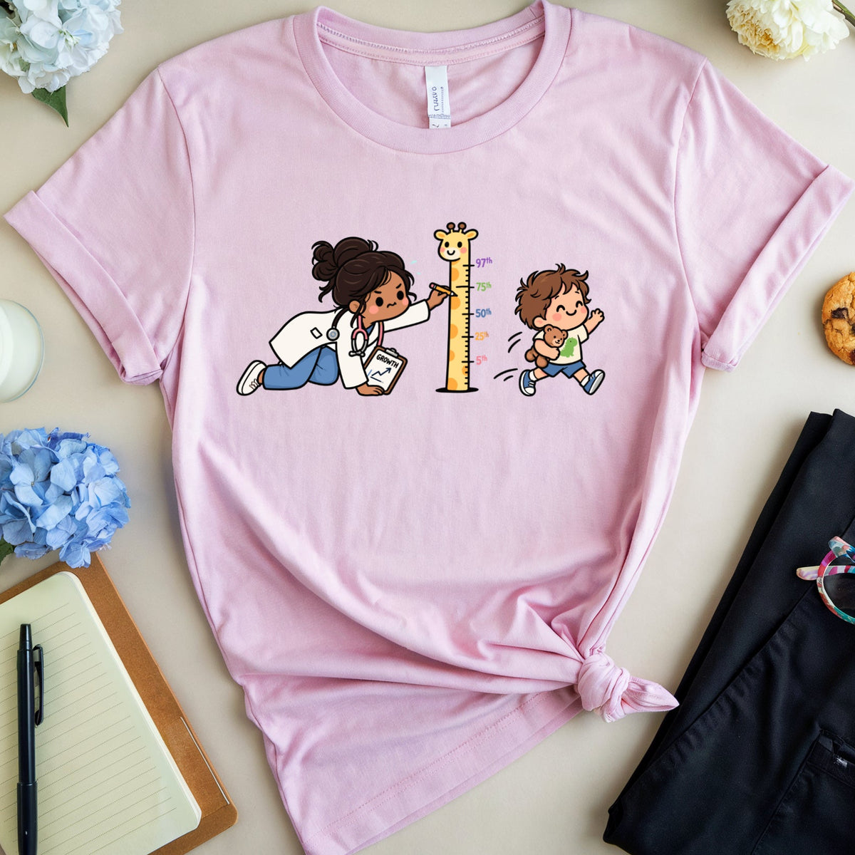 A light pink graphic t-shirt is styled in a flat lay on a beige surface. The t-shirt features a cartoon illustration of a doctor helping a child climb a height chart.