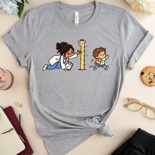 A heather gray graphic t-shirt laid flat on a light beige background, styled with a notebook, glasses, a cookie, a drink, and flowers. The t-shirt features a cartoon graphic of a doctor in a white coat measuring the height of a giraffe while a child watches.