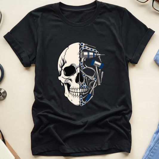 A black short-sleeve T-shirt laid flat on a light beige surface, featuring a large graphic of a human skull split down the middle with one half rendered in white and the other in metallic blue and silver mechanical parts. Surrounding the shirt are a stethoscope, eyeglasses, a notebook with pen, and light blue medical scrubs.